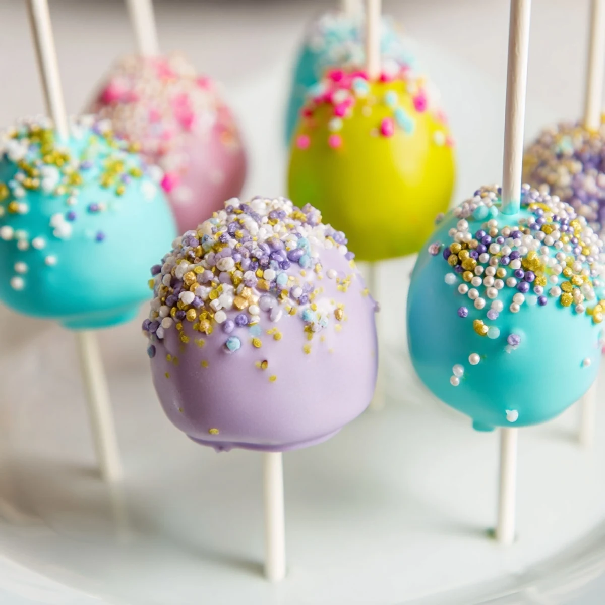 A batch of homemade Easter Egg Cake Pops arranged on a white plate with a few extra sprinkles scattered around for decoration.