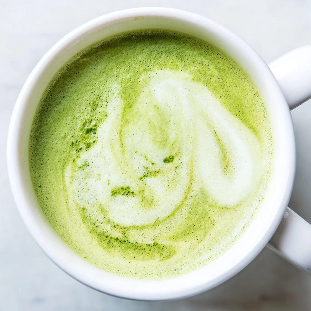 A steaming cup of Green Matcha Latte with Almond Milk sits on a marble countertop, garnished with a light dusting of matcha powder.