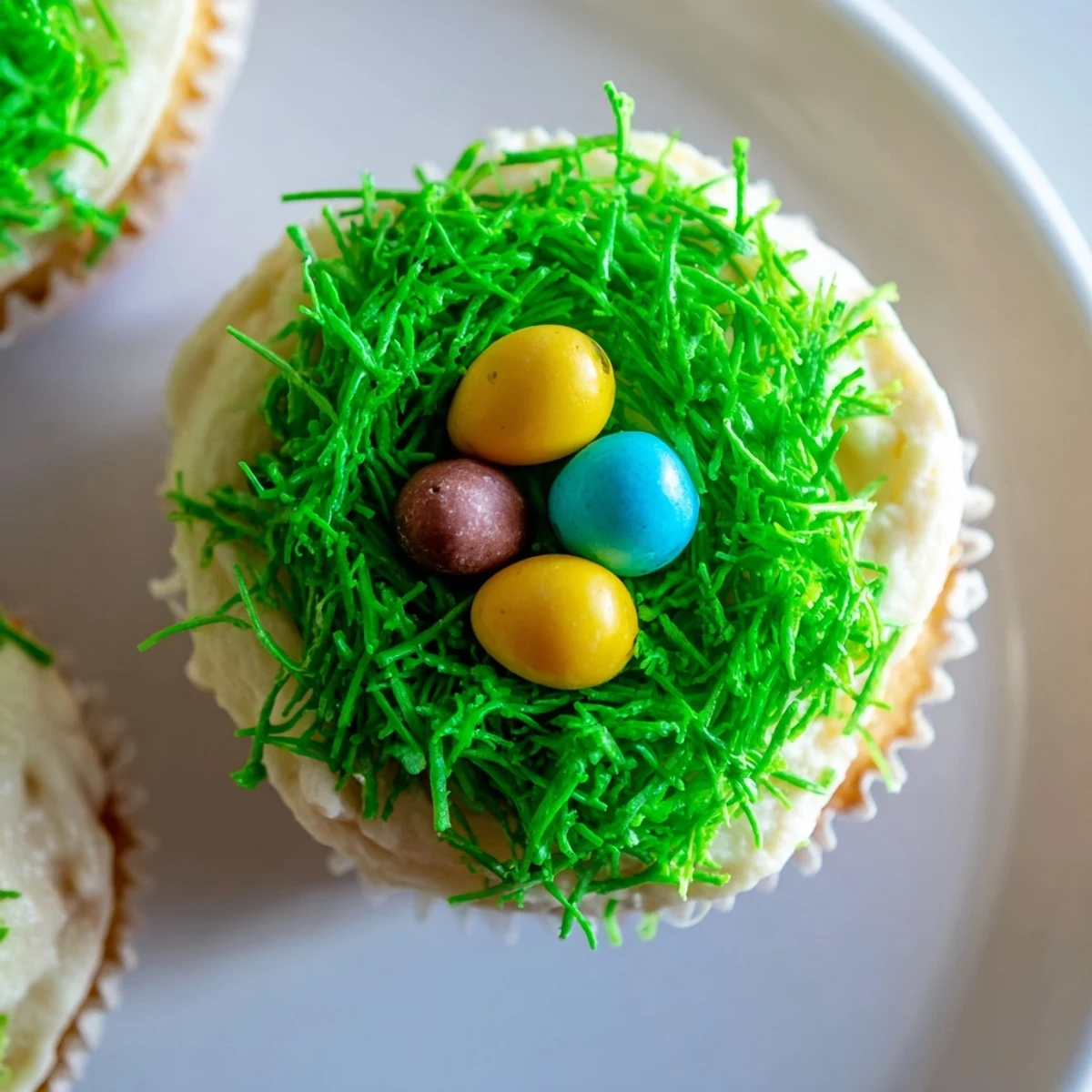 Green-tinted coconut grass and pastel candy eggs top Easter Basket Cupcakes on a wooden board.