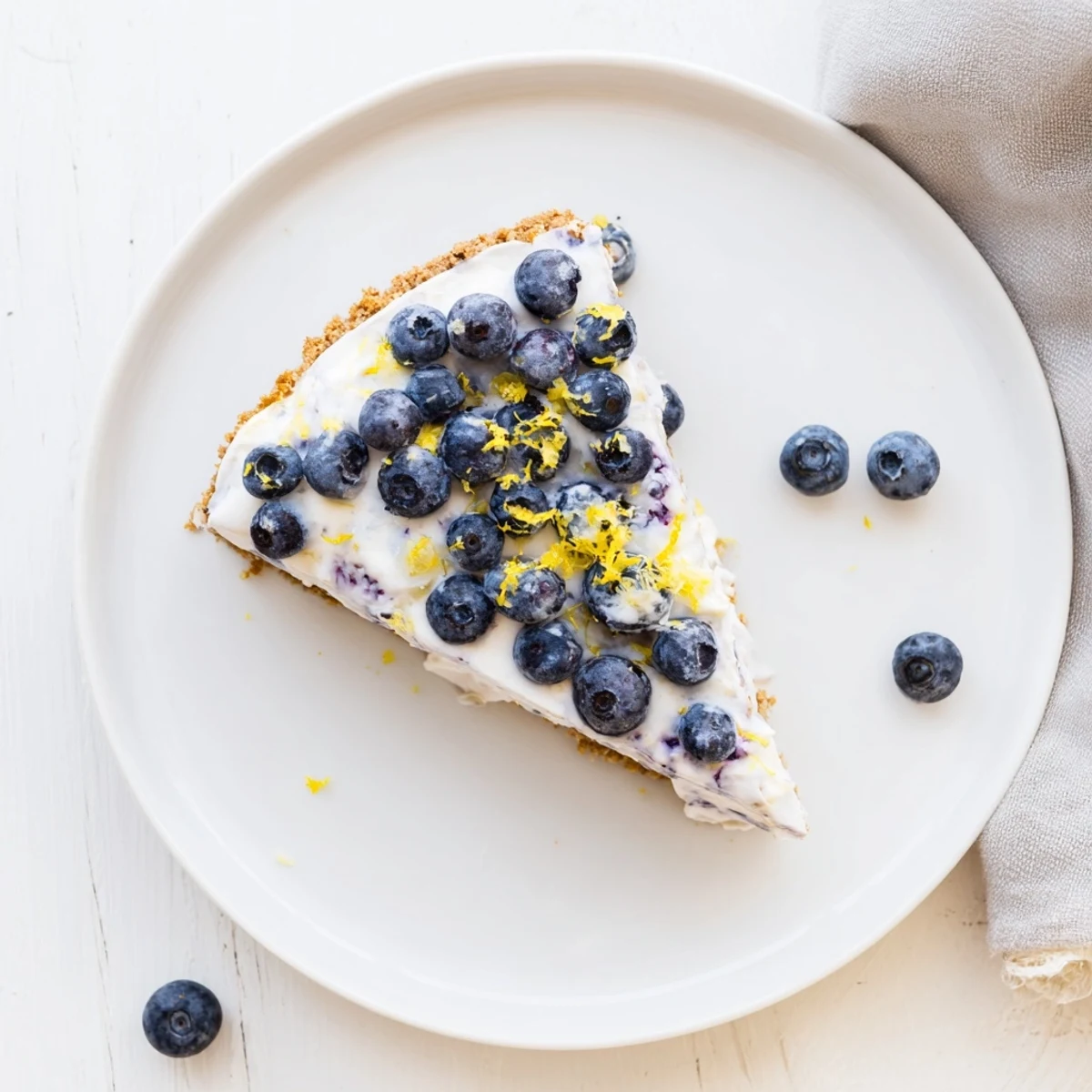 Creamy no bake lemon blueberry cream cake topped with fresh berries and lemon zest on a white plate.