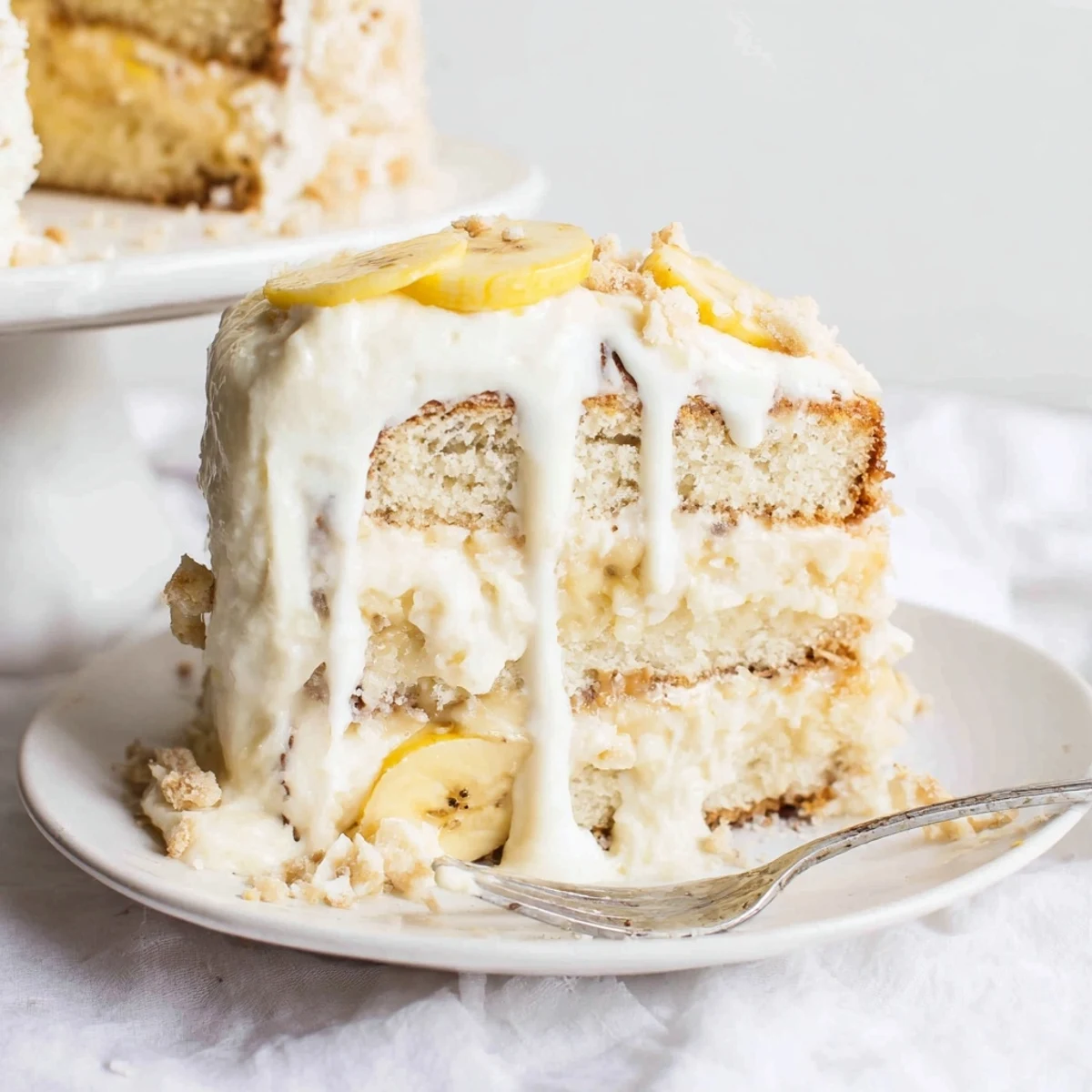 Golden banana pudding cake slice featuring whipped cream topping and fresh banana slices on white plate