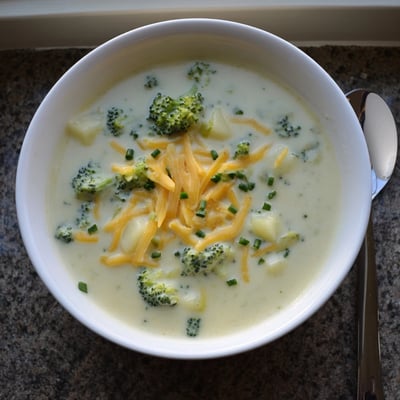 A ladle pours creamy cheesy broccoli potato soup into a bowl with crusty bread on the side.